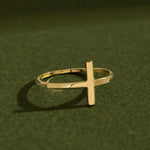 Artistic luxury jewelry photo of a 14K solid yellow gold cross ring placed on textured dark green surface with cinematic lighting and dramatic soft shadows emphasizing the polished gold finish.