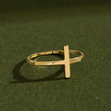 Artistic luxury jewelry photo of a 14K solid yellow gold cross ring placed on textured dark green surface with cinematic lighting and dramatic soft shadows emphasizing the polished gold finish.