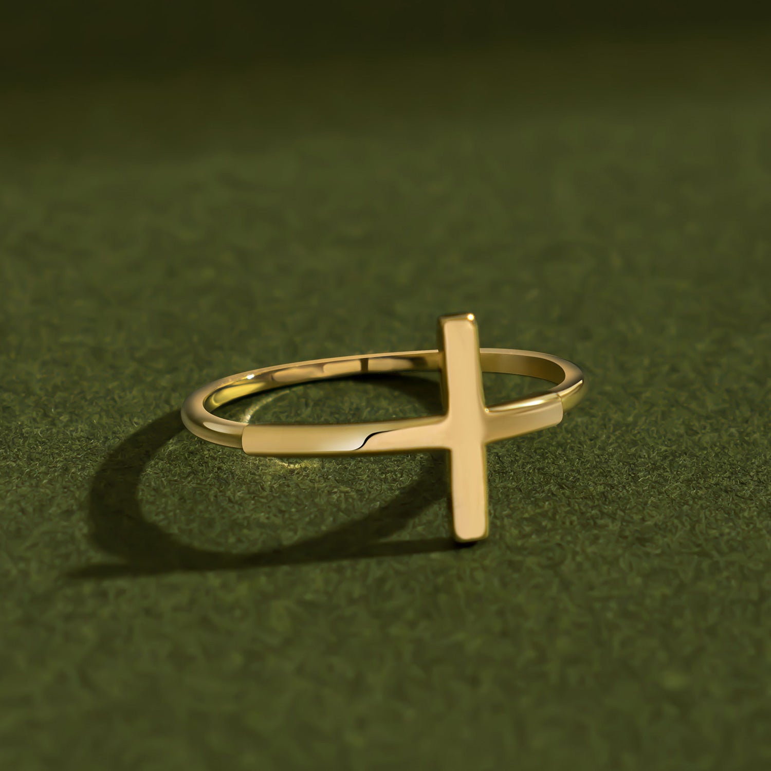 Artistic luxury jewelry photo of a 14K solid yellow gold cross ring placed on textured dark green surface with cinematic lighting and dramatic soft shadows emphasizing the polished gold finish.