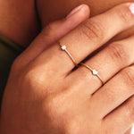 Lifestyle hand photograph showing layered minimalist solid gold rings including a natural diamond birthstone ring paired with a dainty heart ring in elegant stackable styling.