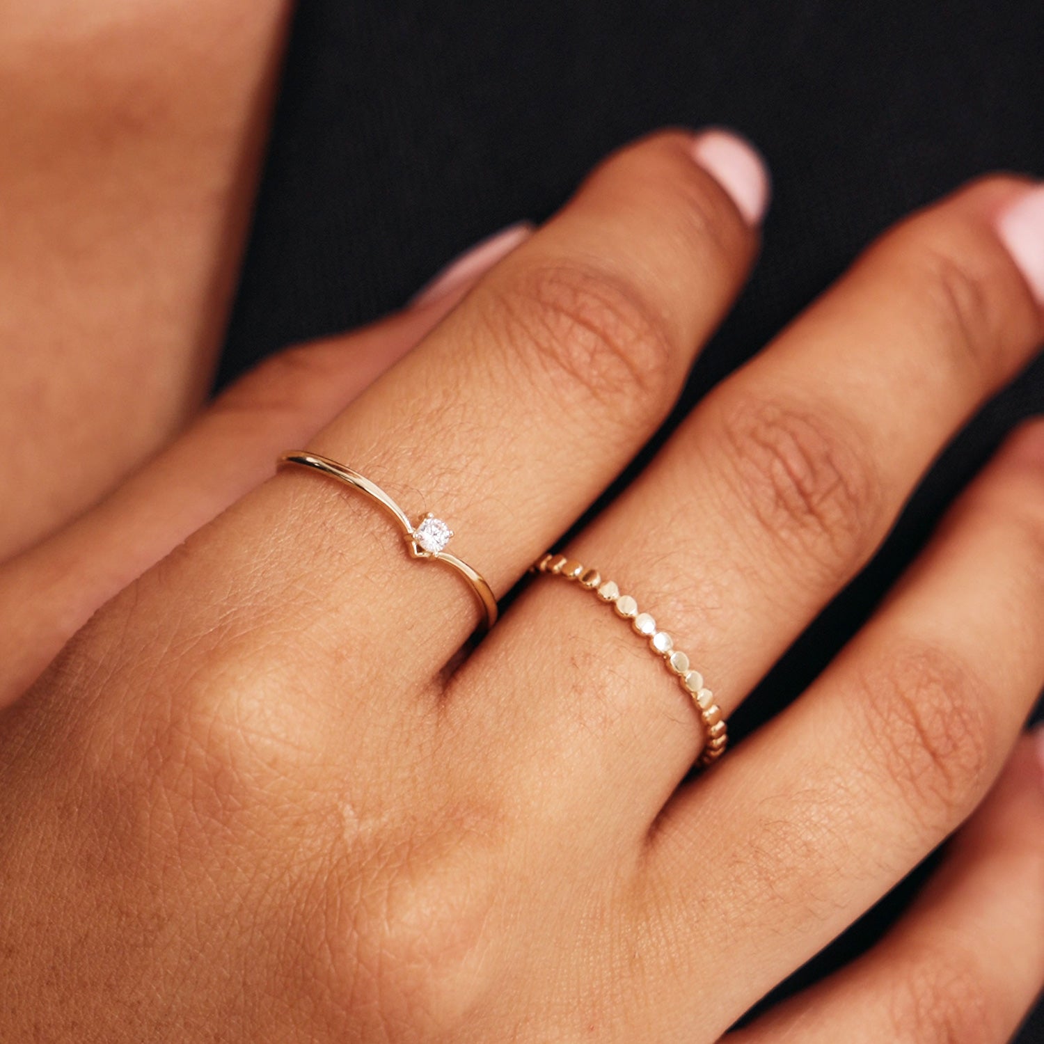 Editorial hand photograph showing layered minimalist 14K solid gold rings including a dainty V shaped diamond promise ring and stackable gold beaded band jewelry.