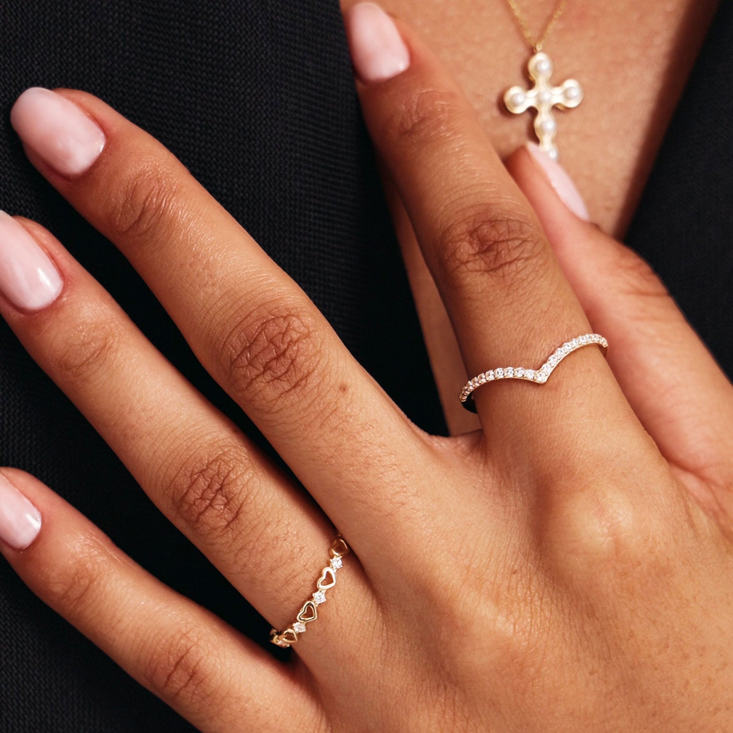 Luxury jewelry styling photograph showing a woman wearing a 14K solid gold diamond wishbone band paired with delicate stackable rings and a gold cross necklace.