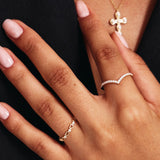 Luxury jewelry styling photograph showing a woman wearing a 14K solid gold diamond wishbone band paired with delicate stackable rings and a gold cross necklace.
